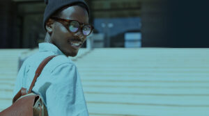 Image of a Black man looking over his shoulder, holding a brown bag and smiling in front of a building