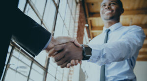 Image of a young Black man who is dressed in a tie and white button down shaking hands with another person