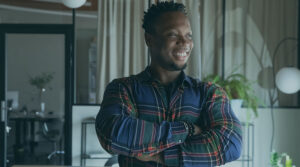 Image of a Black man wearing a plaid shirt crossing his arms and smiling