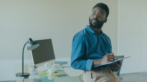 Image of a young professional leaning up against a desk and holding a notepad and pen