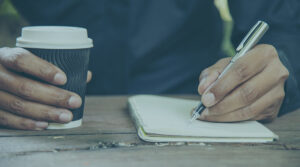 Image of a person holding a pen and writing in a notebook while holding a cup in the other hand