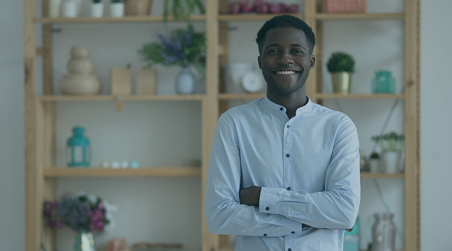 Image of a Black man wearing a light blue button-down with his arms crossed and smiling in front of a shelving unit