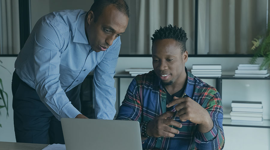A Black man leaning over a desk pointing to something on a laptop screen for another Black man