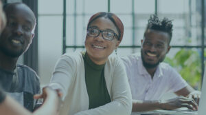 Image of three young Black professionals sitting at a table and smiling as one shakes hands with another person