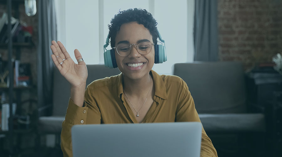 Image of a Black female professional sitting down, wearing headphones and facing a laptop screen while waving