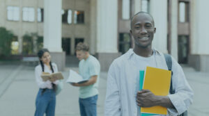 Image of a young Black man standing in front of a building, smiling and holding some books