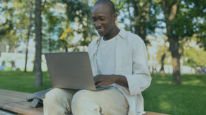 Black student sitting on a bench at a park, smiling, while working on his laptop