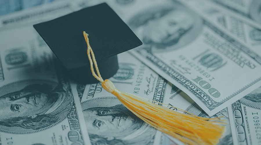 Image of a black graduation cap with a yellow tassel on top of 100-dollar bills