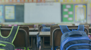 Image of two backpacks strapped on the back of desk chairs in a classroom