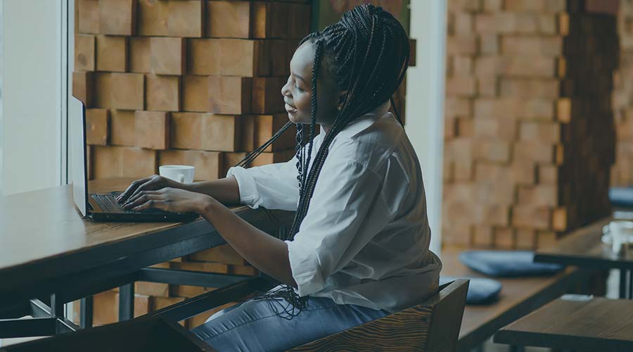 Image of a Black woman sitting on a wood stool and working on her laptop
