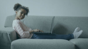 A young Black girl in a pink shirt and jeans sitting on a gray couch with a laptop on her lap