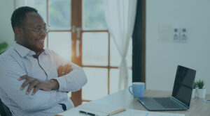 A Black man in a collared shirt and glasses with his arms crossed facing a laptop and desk while smiling