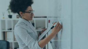 A woman in a striped collared shirt and glasses writing on a whiteboard while holding a clipboard