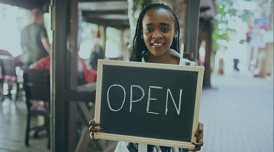 A young black woman holding a chalkboard sign that says “open” outside a business