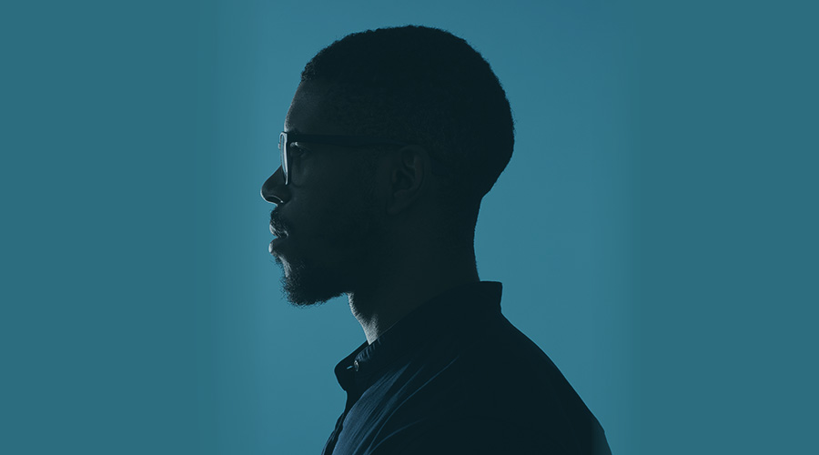 The side profile of a man wearing glasses and a collared shirt against a teal blue background