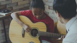 A young girl holds a guitar as she plays while another man listens