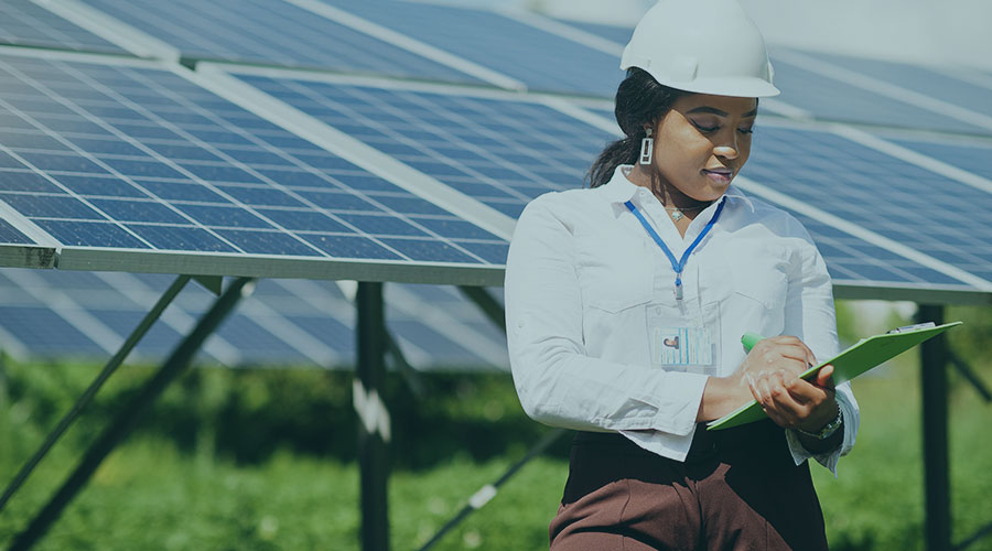 A Black woman wearing a hard hat and holding a clipboard stands in front of solar panels