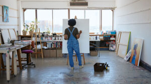 Artist holding a palette standing in front of a white canvas on an easel in a studio
