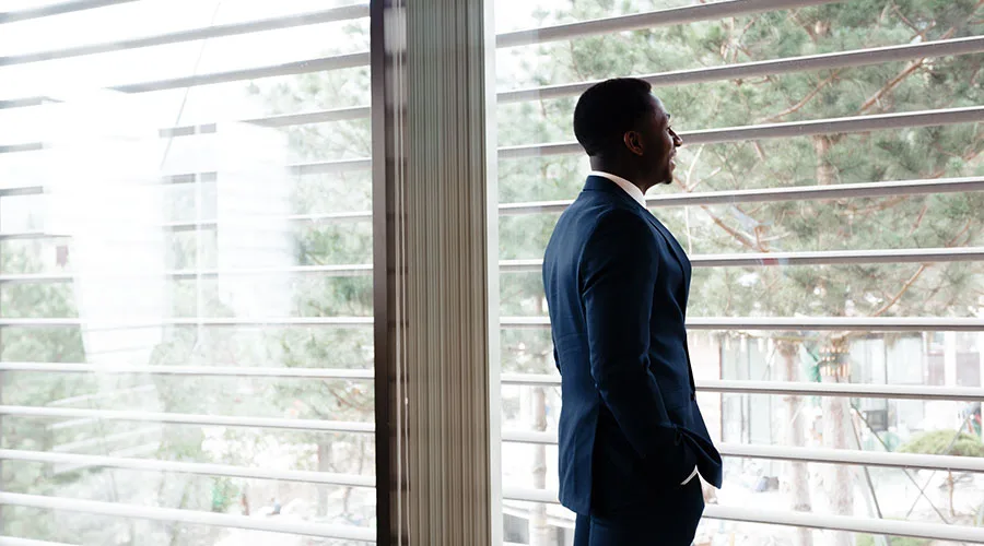 A man wearing a blue suit stands in front of a window while smiling and looking outside