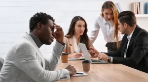 A group of corporate professionals at a table, with three individuals speaking to one another closely, while one sits across the table looking upset.