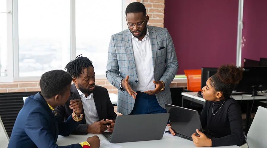 A group of professionals sitting and standing around a conference table in an office setting looking at laptops