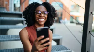 Young Black woman scrolling through her smartphone while sitting on stairs with a city background.