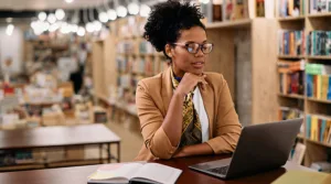 A woman sitting at a table wearing a tan blazer looking at her laptop in a library