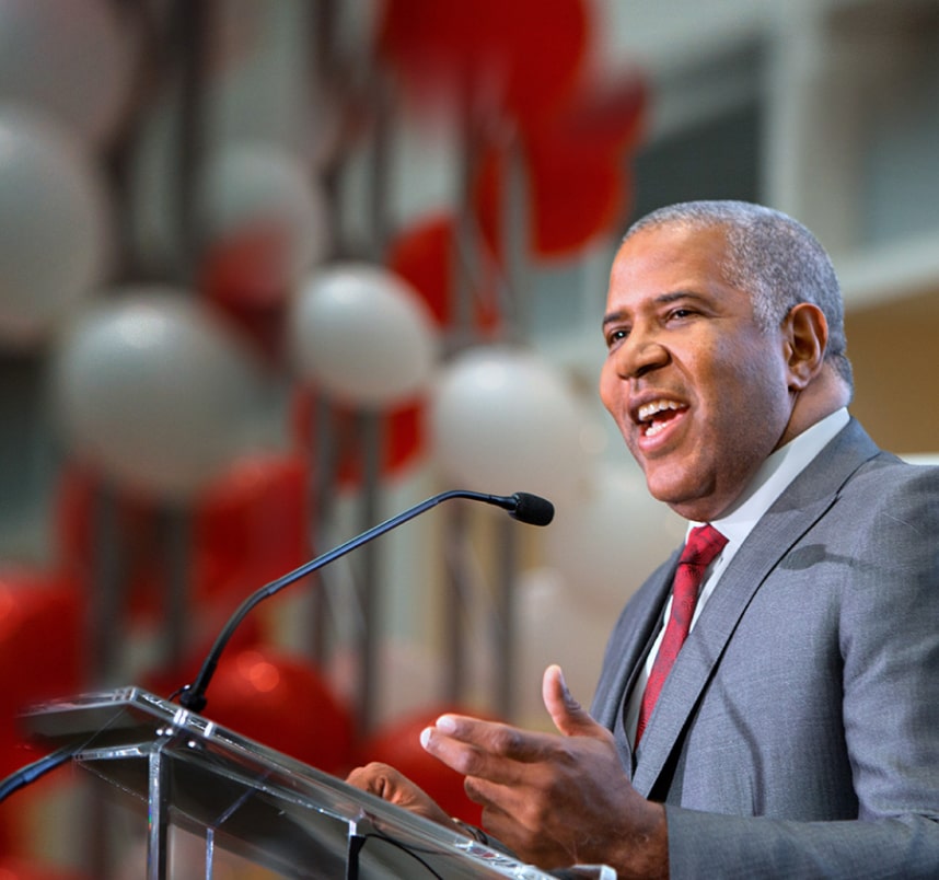 Robert F. Smith wears a suit and delivers a speech at a podium with red and white balloons in the background