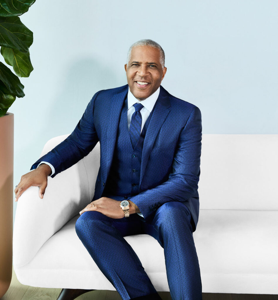 Robert F. Smith in a blue suit, blue tie and white collared shirt sitting on a white couch