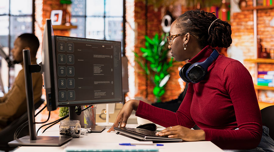 Woman in a red turtle neck and glasses facing two monitors while sitting at a desk.