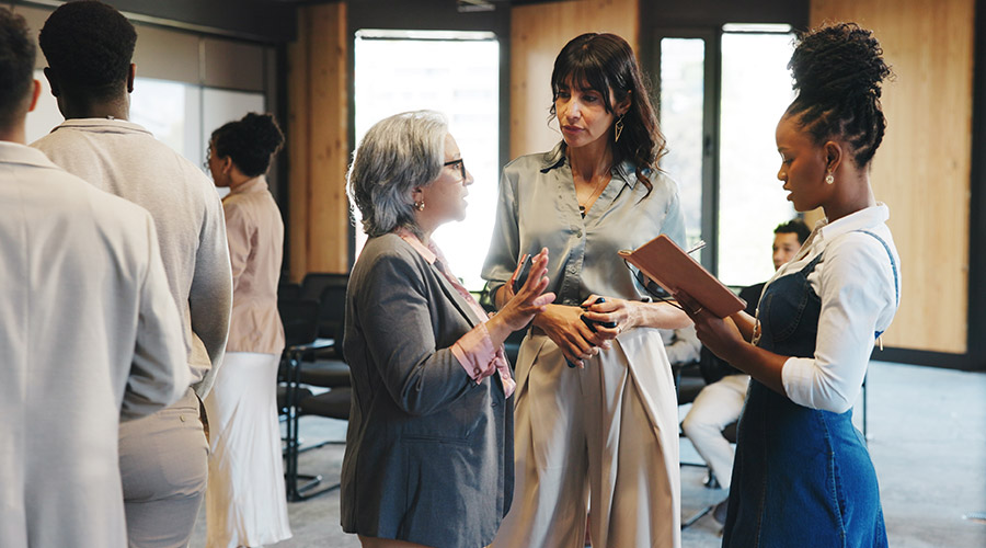 Three professionally dressed women stand in a circle in a conference room, with other people nearby.